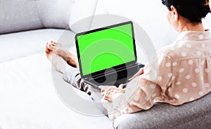 Middle-aged brunette woman on the gray sofa working on green screen laptop, soft focus, stay at home concept, cozy background