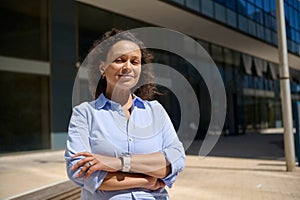 Middle aged business woman standing with her arms folded against office buildings background, smiling, looking at camera
