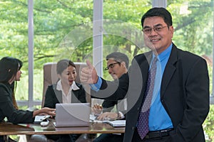 Middle aged businessman holding Thumbs up and standing. Confident senior businessman with group of business people meeting