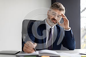 Middle Aged Businessman Sitting At Desk In Office And Writing In Notepad