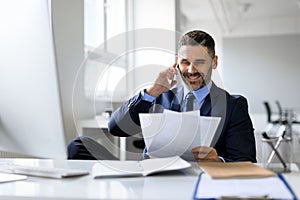 Middle aged businessman talking on cellphone and checking papers, sitting at workplace in office, free space