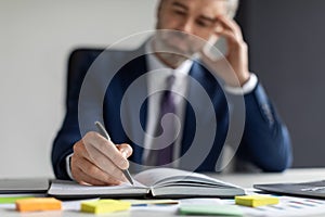 Middle Aged Businessman Writing In Notepad While Sitting At Desk In Office