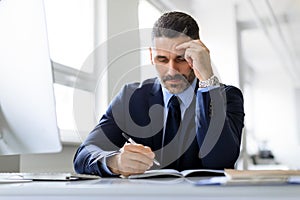 Middle aged businessman writing in notepad, sitting at desk in office interior, free space