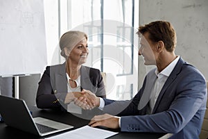 Middle-aged businesspeople sit at desk shake hands express respect