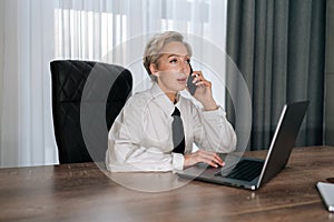 middle-aged businesswoman with blonde hair talking on smartphone siting at office desk with laptop, share helpful