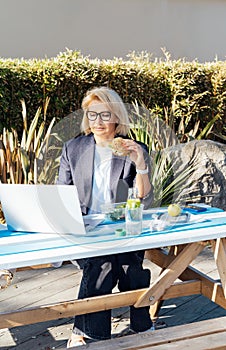 Middle-aged businesswoman having lunch at the park outdoor cafe, picnic area and working on laptop during her break
