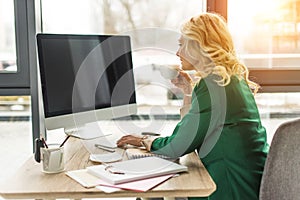 middle aged businesswoman using desktop computer with blank screen and drinking coffee