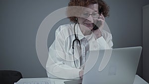 Middle-aged Caucasian female doctor sitting in her consulting room using phone and working on laptop. Elderly general