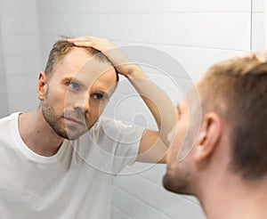 Middle aged caucasian white man with a short beard looks at his hair in the mirror in the bathroom and worried about balding. The