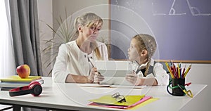 Middle aged caucasian woman and little schoolgirl sitting at the table with tablet and learning math. Education