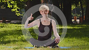 Middle-aged caucasian woman mindfulness slender lady trainer sits on mat on grass lawn in lotus position meditates