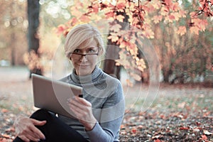 Middle aged caucasian woman sits alone at golden autumn park with tablet, smiling. Casual wear, glasses. Precious age concept.