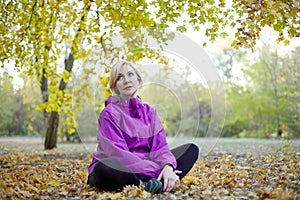 Middle aged caucasian woman sits alone under the big tree at autumn park. Bright casual wear, white wireless earphones, meditation