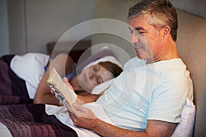 Middle Aged Couple In Bed Together With Man Reading Book