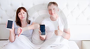 Middle aged couple holding blank screen mobile phone and lying on bed in hotel room bedroom