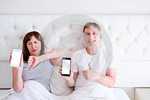 Middle aged couple holding blank screen mobile phone and lying on bed in hotel room bedroom