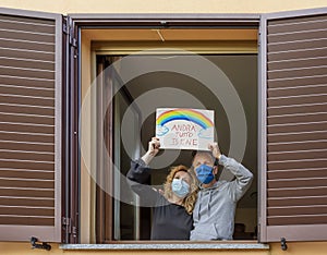 A middle-aged couple looks out the window with protective masks and exposes the sign `Andra` tutto bene`