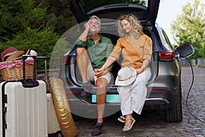 Middle-aged couple sitting in trunk while waiting for charging car before travelling on summer holiday.