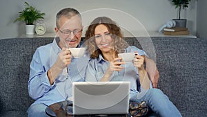 A middle-aged couple watches a movie on their laptop and drinks coffee.