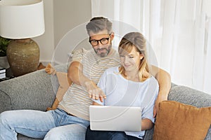 Middle-aged couple wearing casual clothes and sitting at home on the sofa and using a laptop