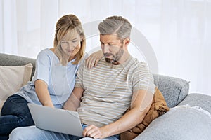 Middle-aged couple wearing casual clothes and sitting at home on the sofa and using a laptop