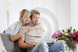 Middle-aged couple wearing casual clothes and sitting at home on the sofa and using a laptop