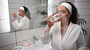 Middle aged dark-haired African American woman in white waffle bathrobe drinking lemon water standing in the bathroom and smiling