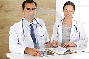 Middle aged doctor man filling up medical documents or prescription with his female colleague. Group of doctors at work