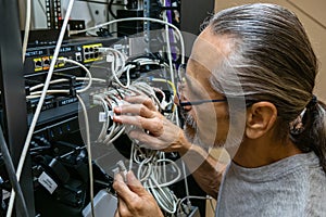 A middle-aged engineer with glasses and a ponytail plugs in network cables at the back of a rack