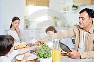 Middle aged father enjoying meal together with his family, using digital tablet while sitting at the table in kitchen at