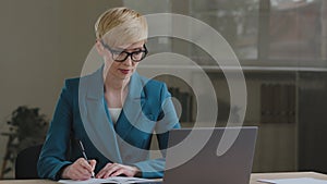 Middle aged female entrepreneur wears glasses using laptop writing notes sitting at office table adult 40s business