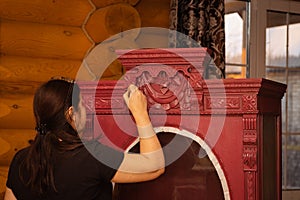 Middle aged female patinating beautiful carved ornaments on cupboard made of wood with brush in hand in workshop for