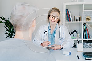 Middle aged female professional doctor physician consulting gray hair patient, filling form at consultation, and talking
