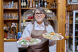 Middle aged female restaurant owner in apron with plates of cooked food