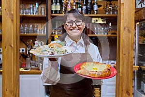 Middle aged female restaurant owner in apron with plates of cooked food