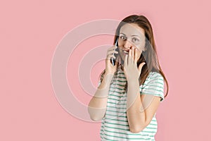 A middle-aged girl in casual clothes is talking on the phone and covers her mouth with her palm in surprise. Studio photo on a