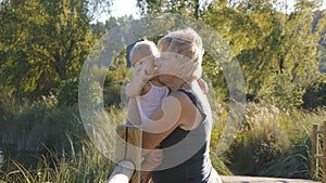 Middle aged grandmother holds her blonde baby girl standing on wooden bridge in natural park