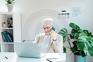 Middle aged gray hair businessman talking on mobile phone and using laptop while working at home office. Confident