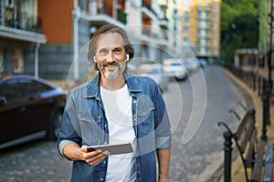 Middle aged grey haired freelancer man hold digital tablet in hands having a call while standing on urban streets