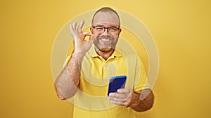 Middle-aged handsome caucasian man smiling while holding cellphone, messaging with ok gesture against yellow isolated background
