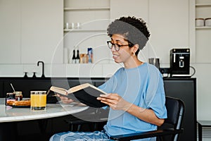 Middle-aged happy woman reading book while having breakfast at home