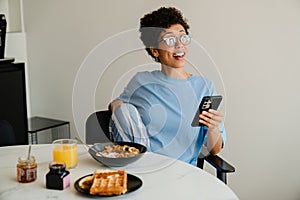 Middle-aged happy woman using cellphone while having breakfast at home