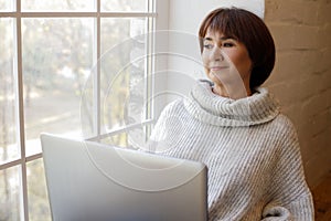 Middle aged happy woman working or learning on her laptop sitting near window in quarantine lockdown. Social distancing Self