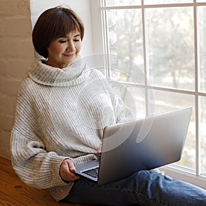 Middle aged happy woman working or learning on her laptop sitting near window in quarantine lockdown. Social distancing Self