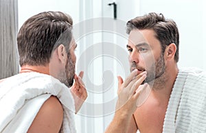 Middle aged hispanic man looking in mirror, facial skin and stubble. Male beauty care product. Skincare, home spa