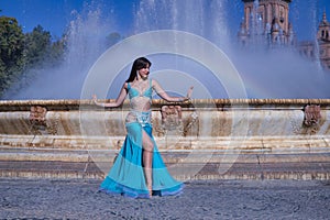 Middle-aged Hispanic woman, with turquoise costume and rhinestones, for belly dancing, dancing in front of a water fountain where