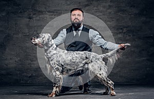 Middle-aged hunter dressed in elegant clothes sits on his knee and shows the full length of his purebred English Setter.