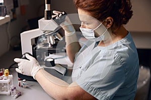 Middle aged lab technician adjusts microscope to research material sample at workplace