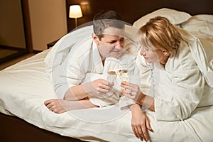 Middle-aged lady and her husband are drinking champagne in bed