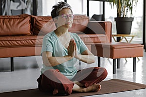 A middle aged lady is practicing yoga at home. A woman with closed eyes and palms, folded together on chest, sits on a